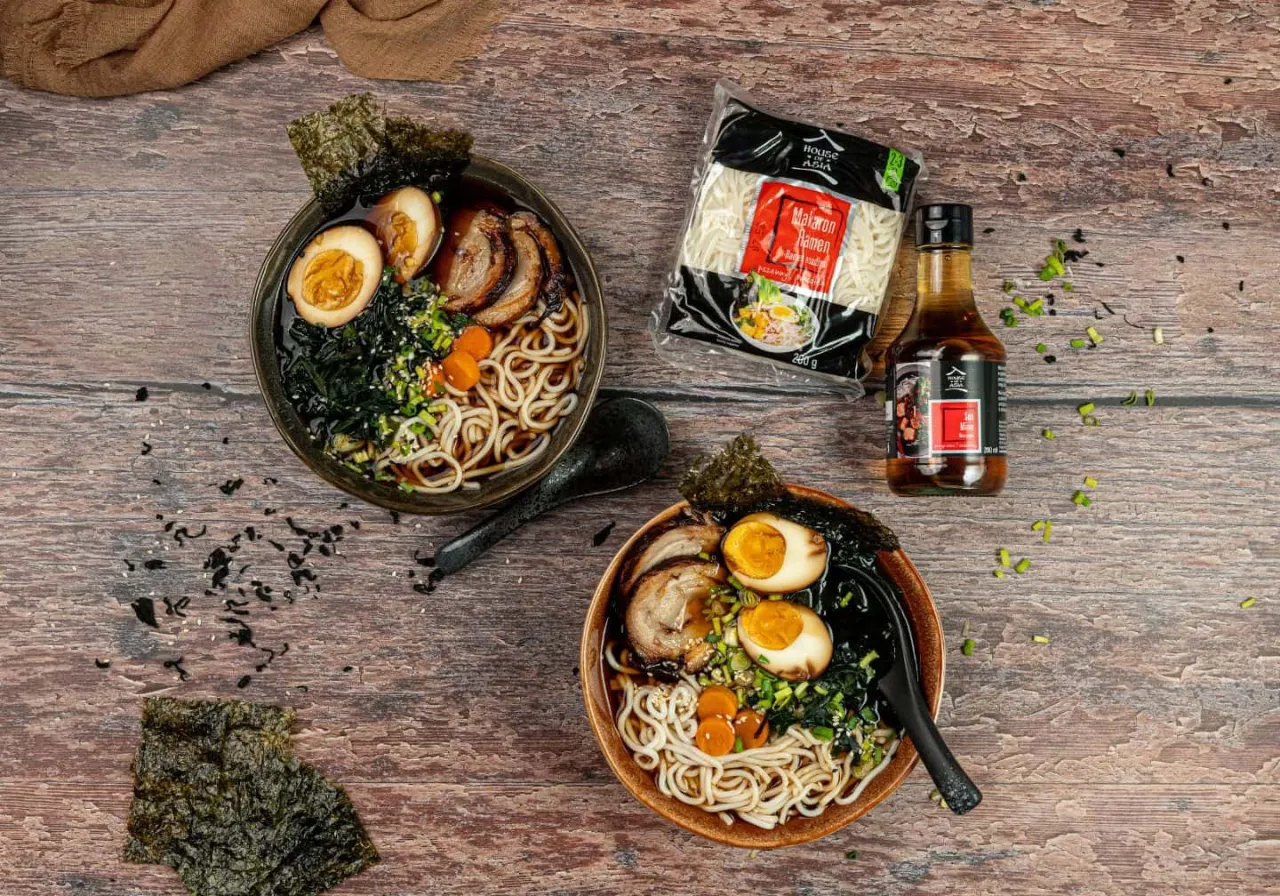 Dwa miski aromatycznego ramenu z jajkiem, mięsem i nori. Obok opakowanie makaronu i sosu, idealne do przygotowania własnego ramenu przepis.