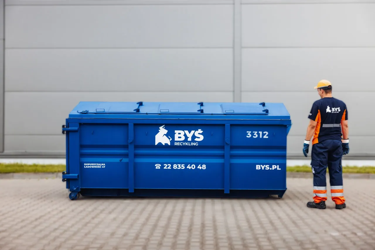 Pracownik firmy BYŚ RECYKLING stoi obok niebieskiego kontenera. Dowiedz się, ile kosztuje kontener na śmieci Warszawa.