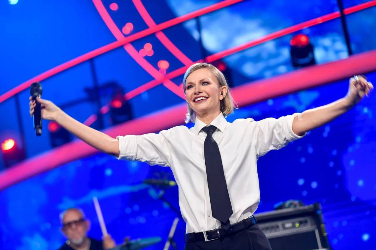 Kto śpiewa orła cień? Wokalistka na scenie z mikrofonem, w białej koszuli i krawacie, z ramionami rozłożonymi szeroko.