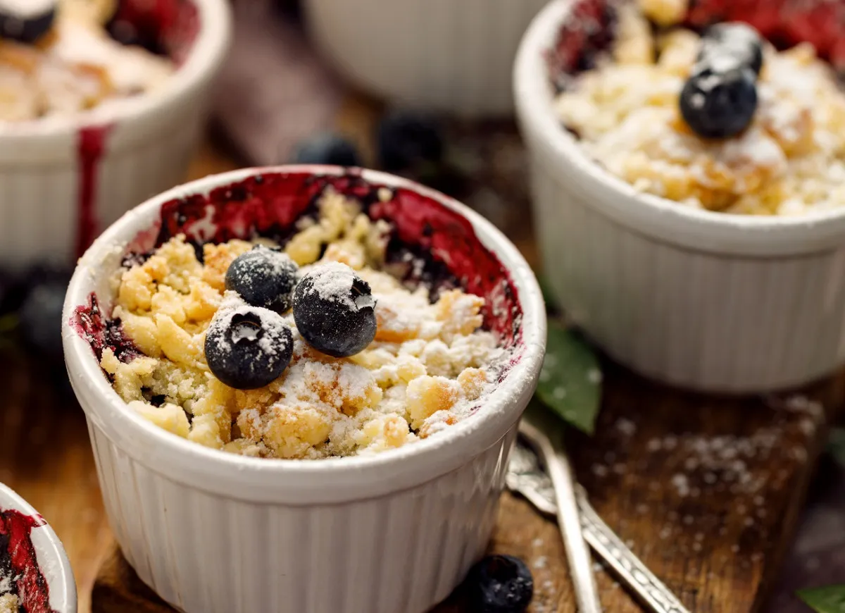 Pyszny biszkopt z jagodami i kruszonką, posypany cukrem pudrem, w białych kokilkach.