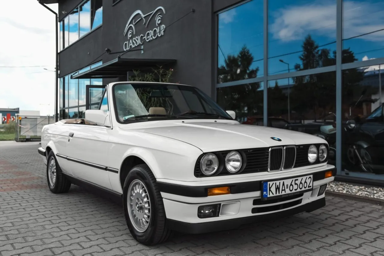 Białe bmw e30 cabrio z beżowym wnętrzem zaparkowane przed budynkiem z napisem 