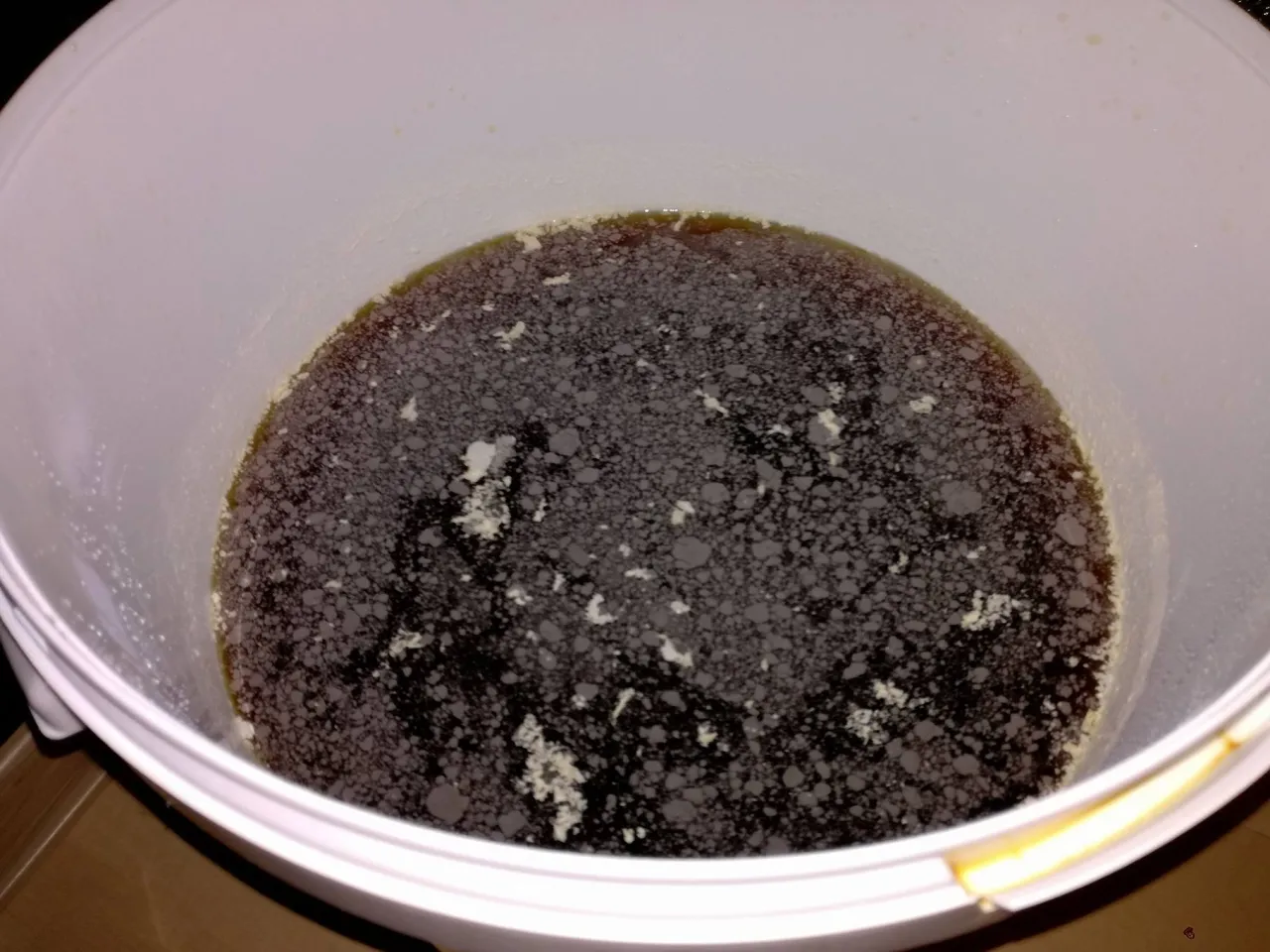 Objawy zakażenia warki: jak rozpoznać pleśń a jak kożuch drożdżowy? Na powierzchni ciemnego płynu w białym wiadrze widać szaro-białe skupiska i drobne pęcherzyki.