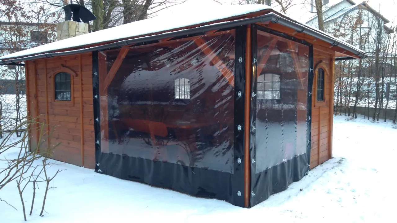 Drewniana altanka z przezroczystymi zasłonami, które pomagają jak zabezpieczyć altankę przed wiatrem. Widać śnieg i zimowy krajobraz.