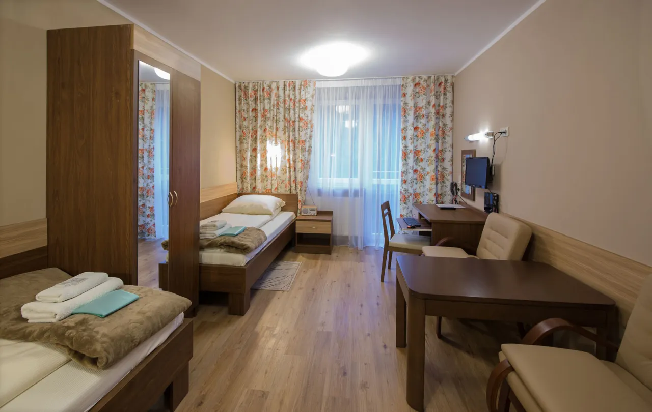 Pok&oacute;j w sanatorium Wł&oacute;kniarz Busko Zdr&oacute;j: dwa ł&oacute;żka, szafa, biurko z komputerem, st&oacute;ł i krzesła.
