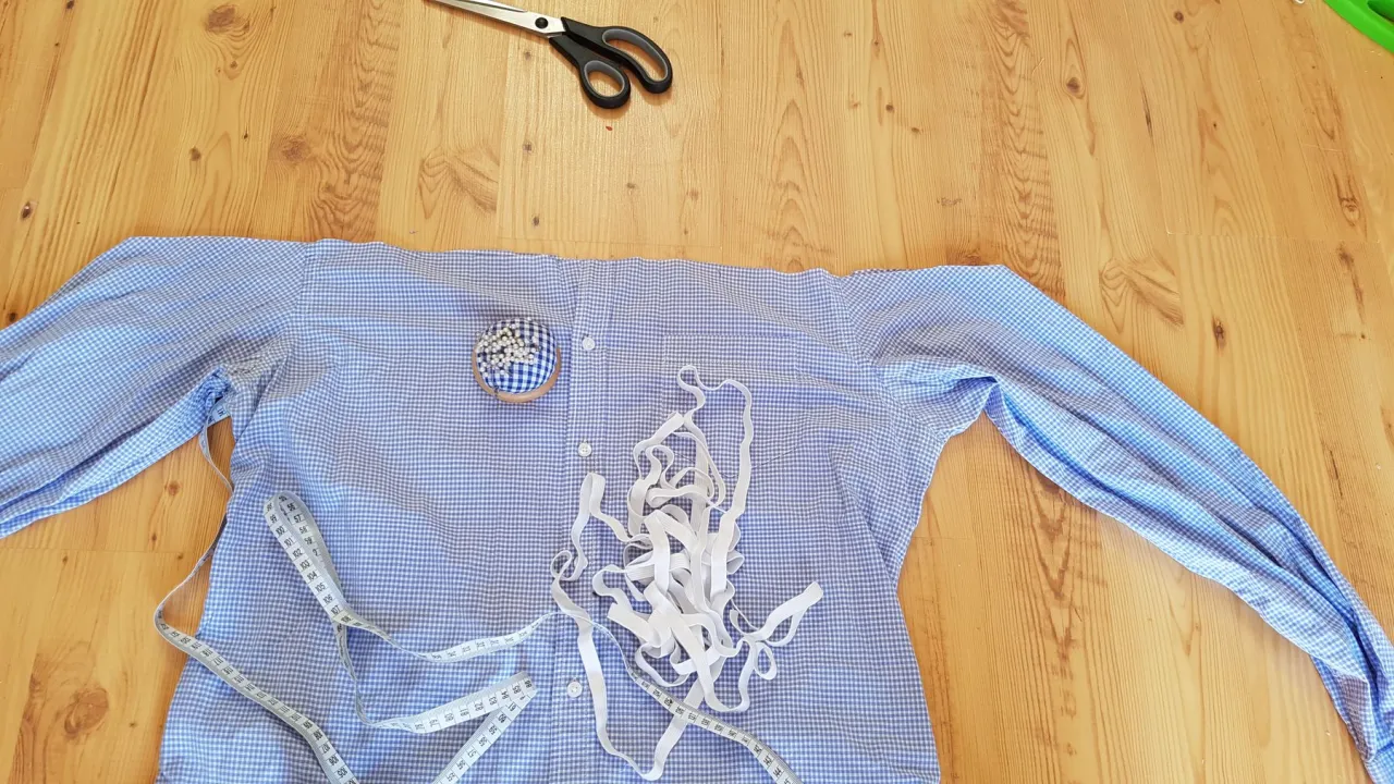 Przygotowania do szycia bluzki hiszpanki: błękitna koszula w kratkę, nożyczki, miarka, agrafka z białymi szpilkami i biała gumka.