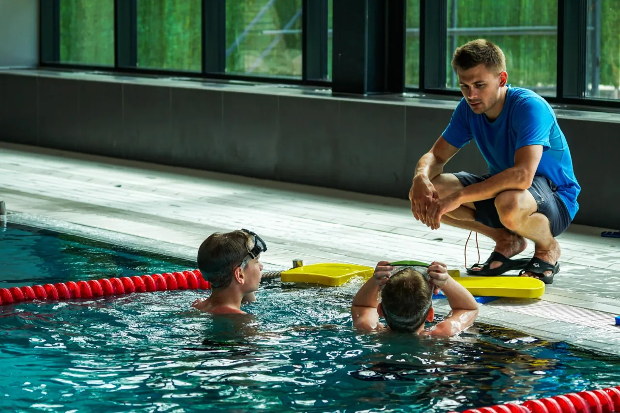 Instruktor pokazuje, jak nauczyć się pływać w starszym wieku. Dzieci w basenie słuchają uważnie.