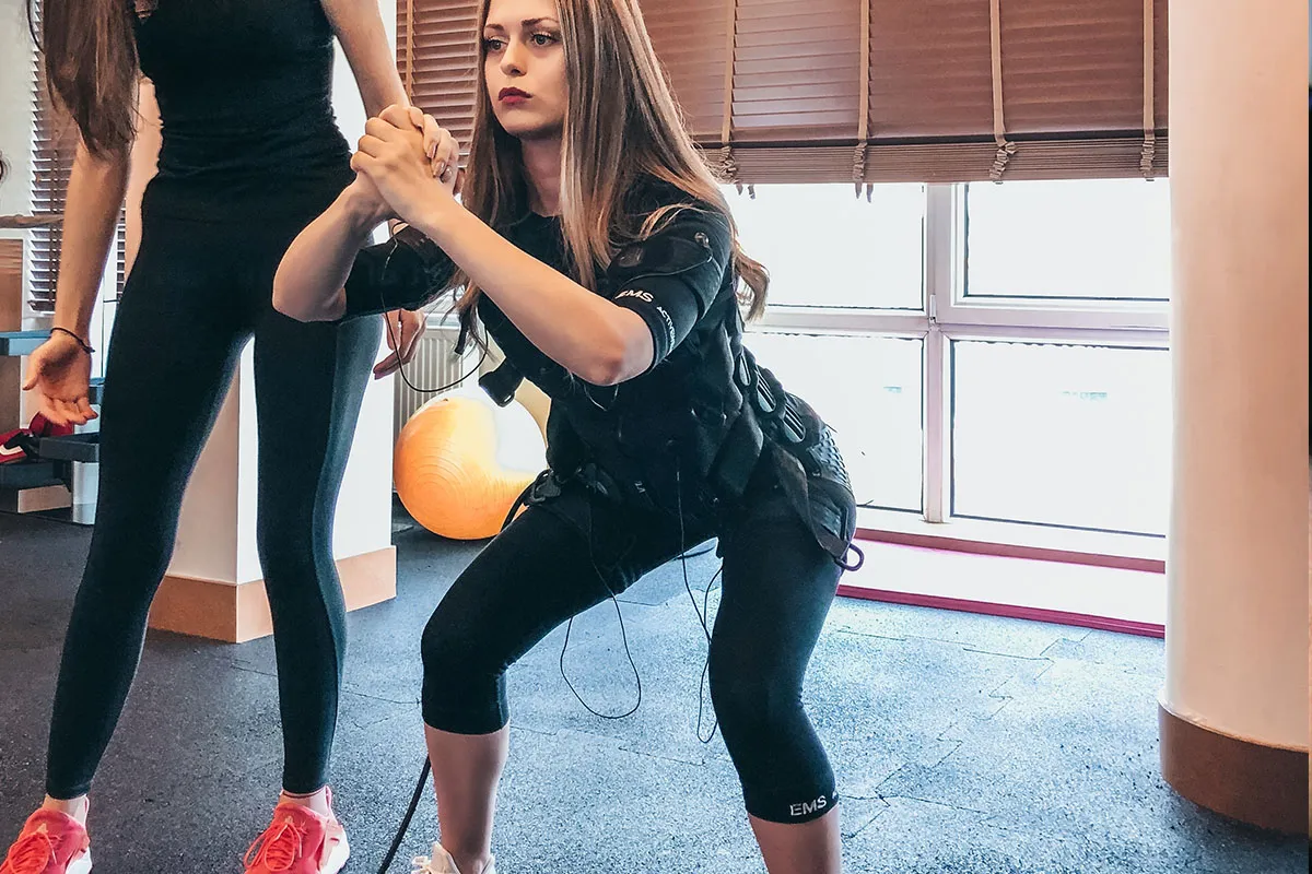 Kobieta w kamizelce EMS wykonuje przysiad, pokazując, na czym polega trening ems. Obok stoi druga osoba.