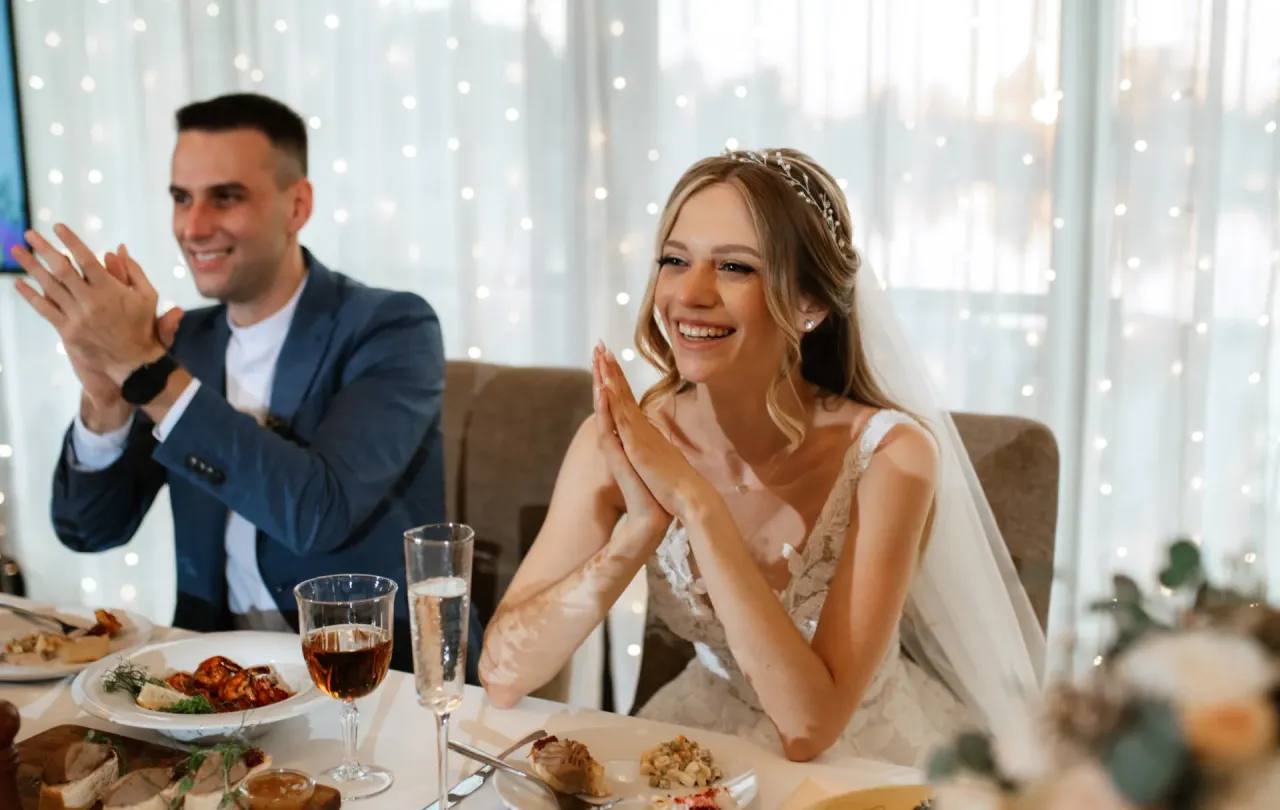 Para młoda cieszy się z kameralnego obiadu weselnego zamiast wesela. Uśmiechnięci, klaszczą przy stole pełnym smakołyków.