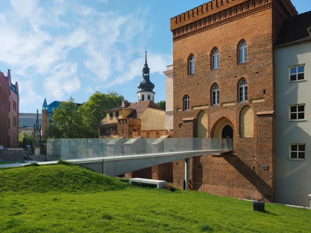 Nowoczesny mostek nad zielonym trawnikiem prowadzi do zabytkowej ceglanej wieży, jednego z miejsc, co można zwiedzić w Opolu. W tle widać wieżę kościelną.