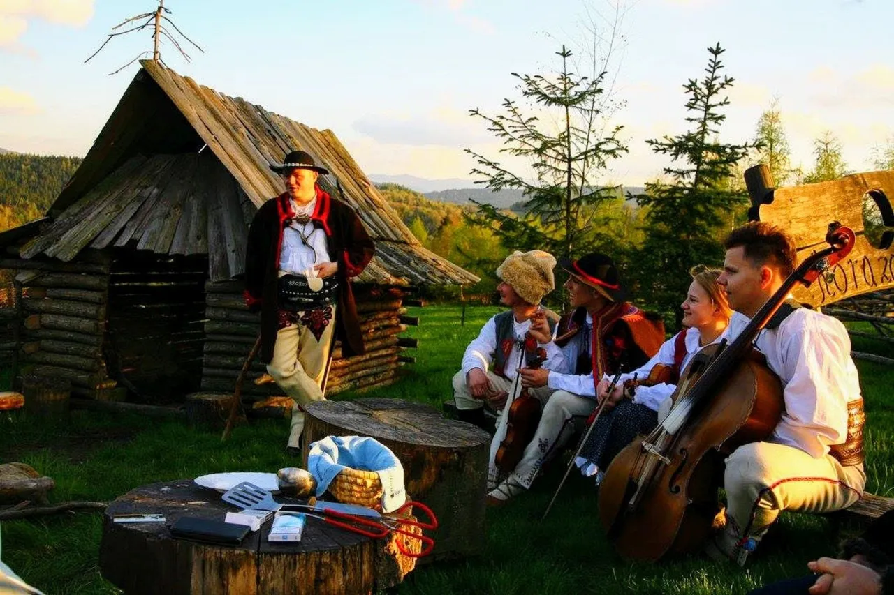 Muzycy w strojach g&oacute;ralskich grają przy bac&oacute;wce. W tle drewniana chata i drzewa.