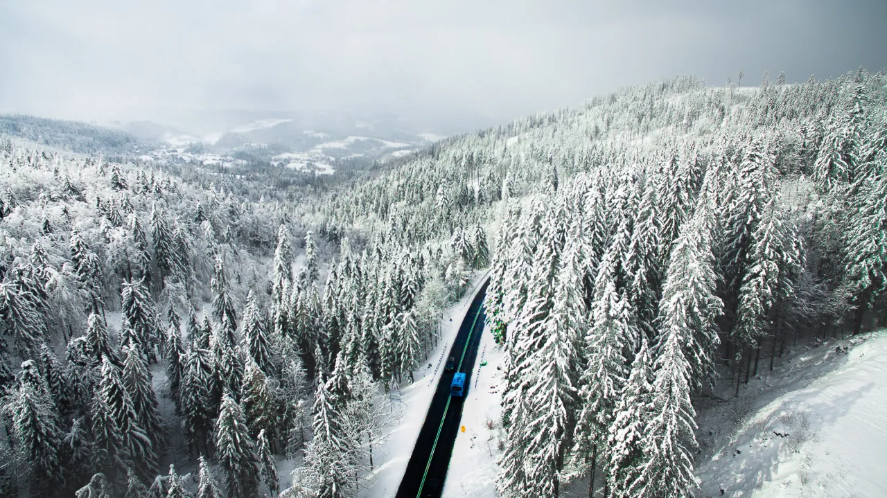 Zimowa droga przez zaśnieżony las w Beskidach. W oddali widać zabudowania, a nad nimi mgliste pasmo g&oacute;rskie.