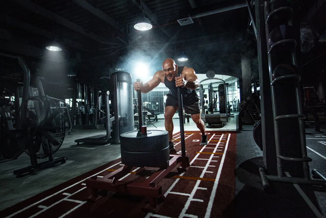 Mężczyzna pcha sanie z obciążeniem na bieżni w siłowni. Pokazuje, jak połączyć crossfit z siłownią, łącząc siłę i wytrzymałość.