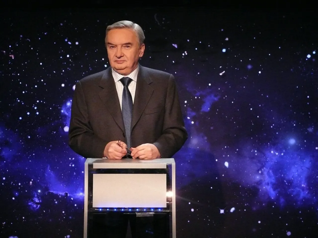 Mężczyzna w garniturze stoi przy m&oacute;wnicy na tle kosmicznego nieba. Jeden z dziesięciu od kiedy zaczął karierę.