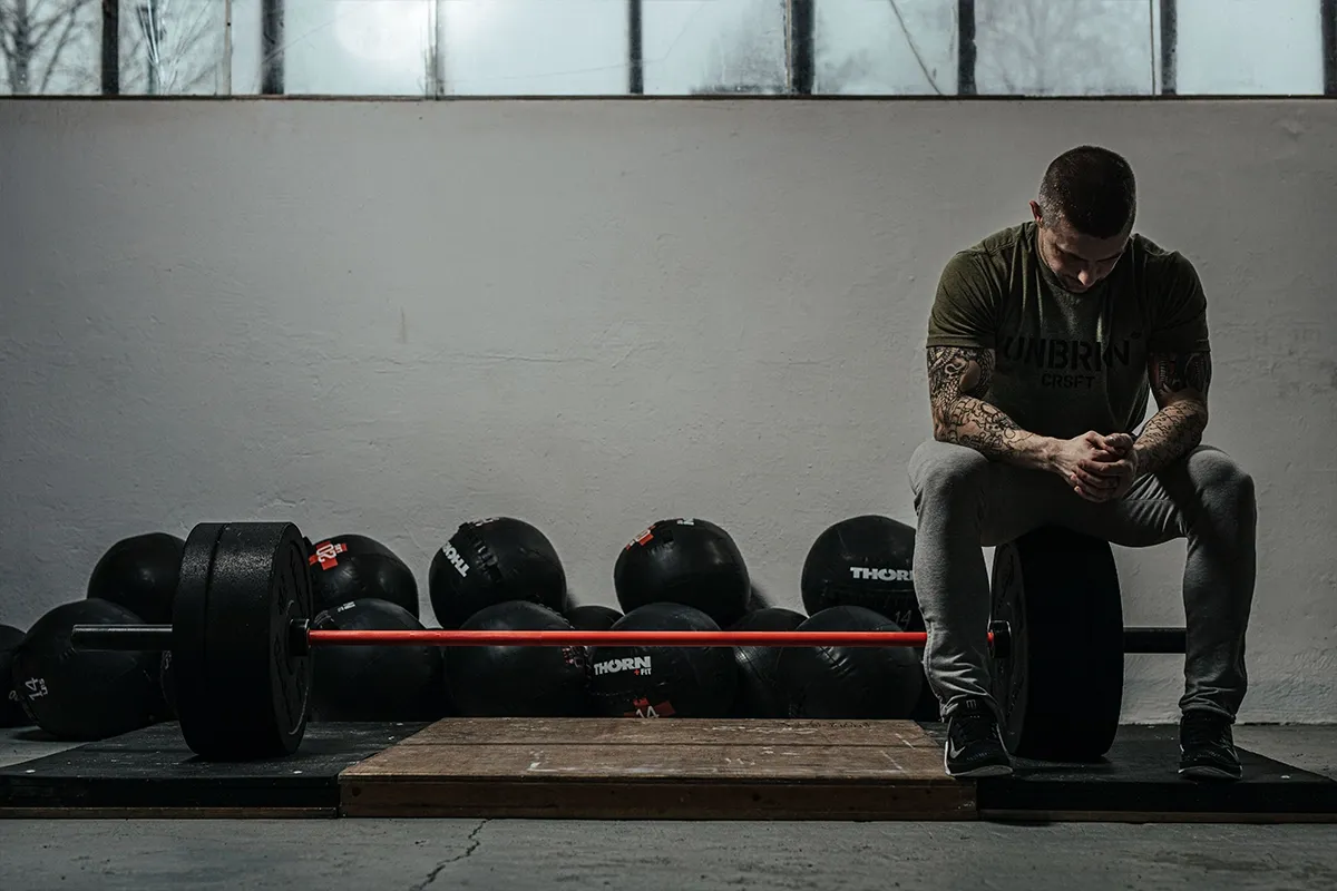 Mężczyzna z tatuażami siedzi na talerzu od sztangi, otoczony piłkami lekarskimi i gryfem. Inspiracja, jak ćwiczyć crossfit w domu.
