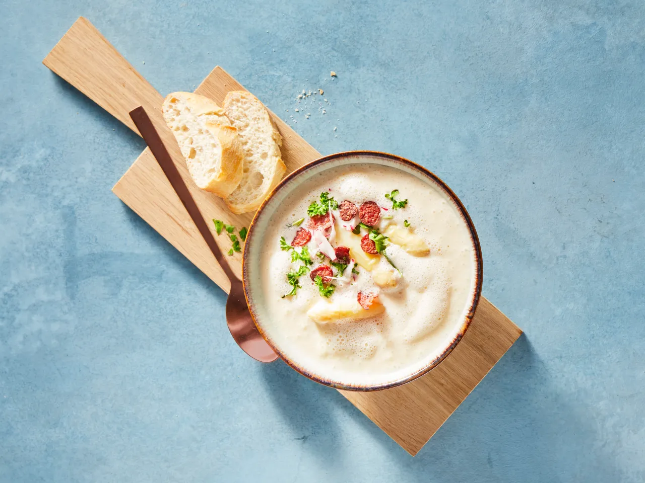 Kremowa zupa z szparagami, kiełbaską i natką pietruszki. Chleb i łyżka na desce. To jak zahartować śmietanę, by zupa była aksamitna.