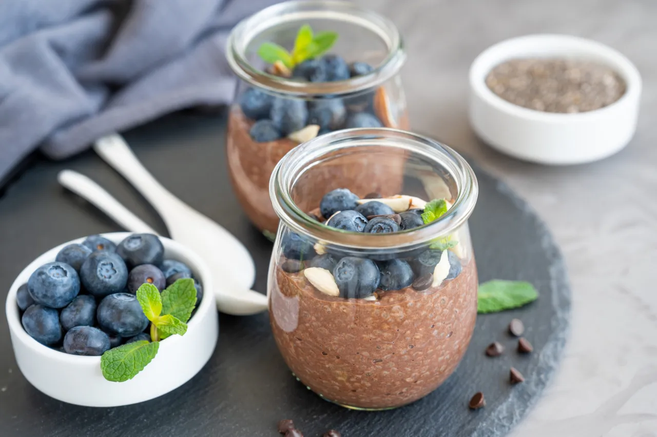 Pyszne śniadanie z chia w słoiczkach, udekorowane bor&oacute;wkami, migdałami i miętą. Obok miseczka świeżych bor&oacute;wek.