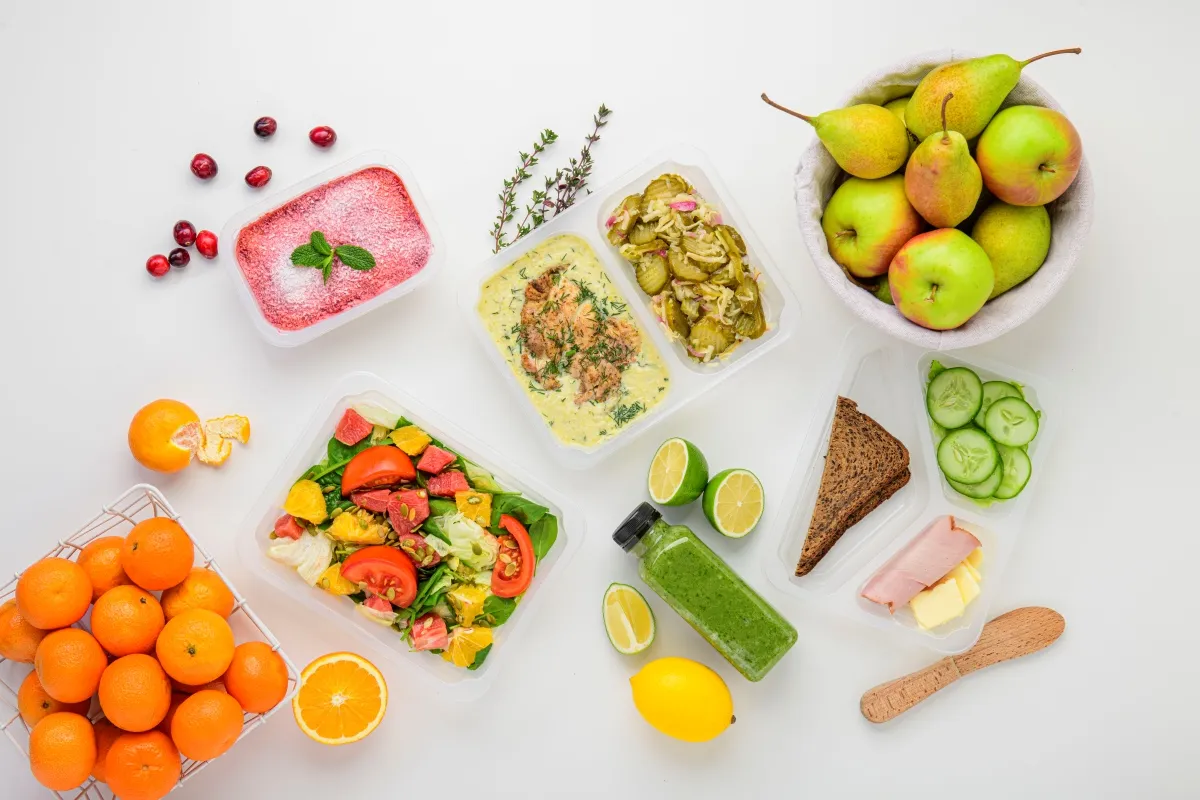 Catering dietetyczny co to? Zestawy posiłków w pojemnikach: sałatka, ryba z sosem, kanapka, owoce, warzywa i zielony koktajl.