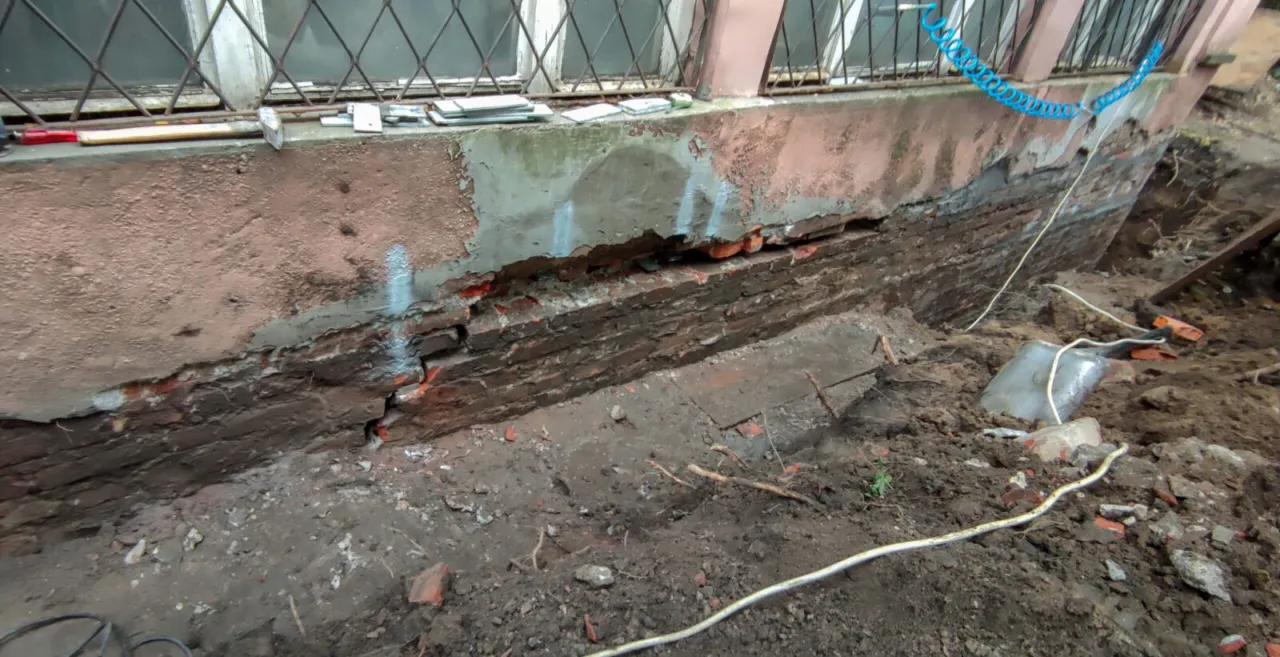 Odsłonięty, pęknięty mur ceglany wymaga prac, by jak wzmocnić fundamenty. Widać narzędzia i ziemię wokół.