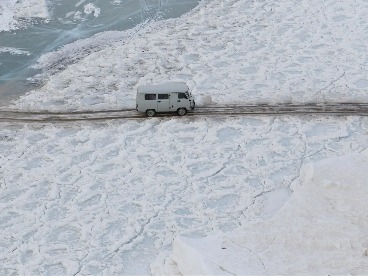 Biały van jedzie po lodzie, jakby kiedy bałtyk zamarzł całkowicie. Widać pęknięcia i nierówności na zamarzniętej tafli.