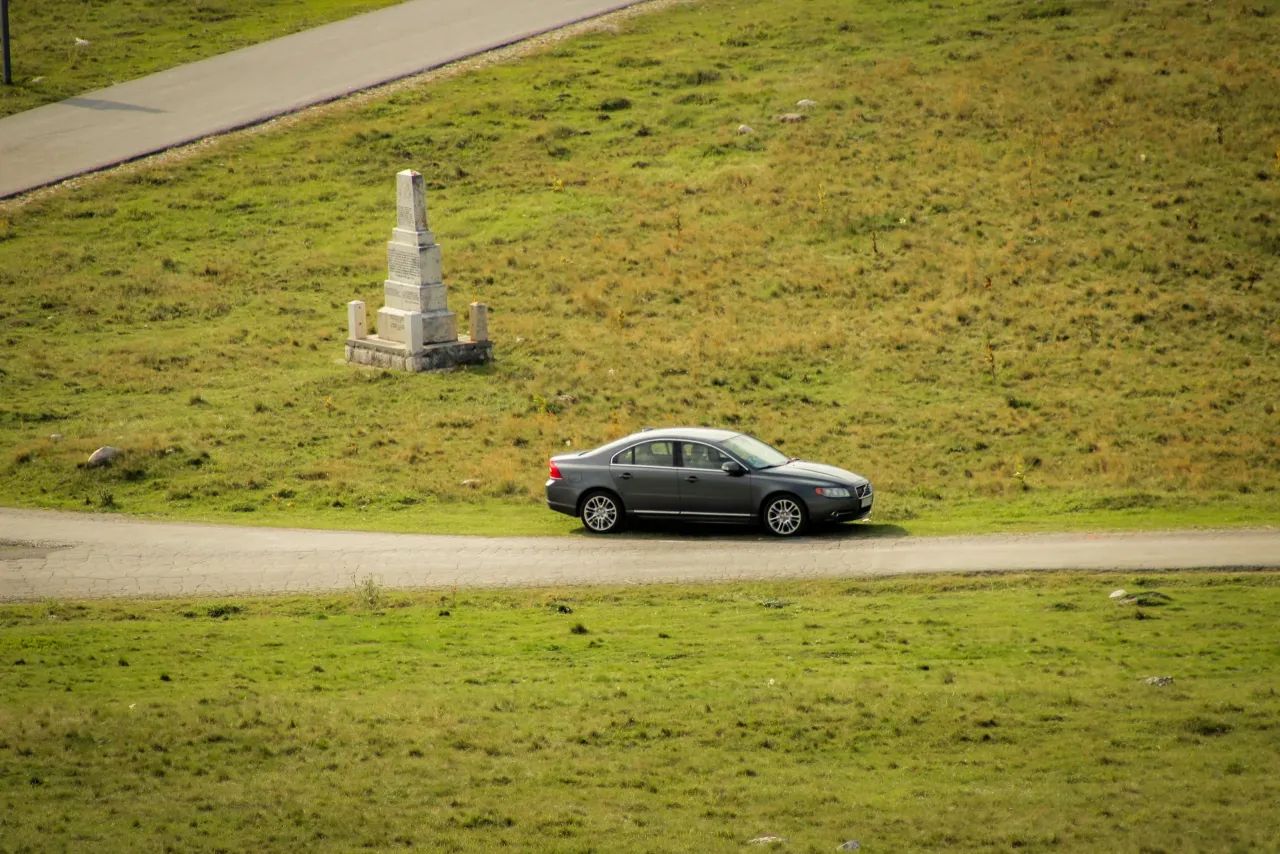 Szary samochód na drodze w Czarnogórze, obok kamiennego pomnika na zielonym wzgórzu.
