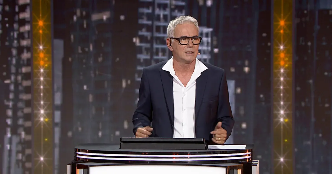 Mężczyzna w okularach i garniturze stoi na scenie. Tło przypomina nocne miasto. To scena z programu 