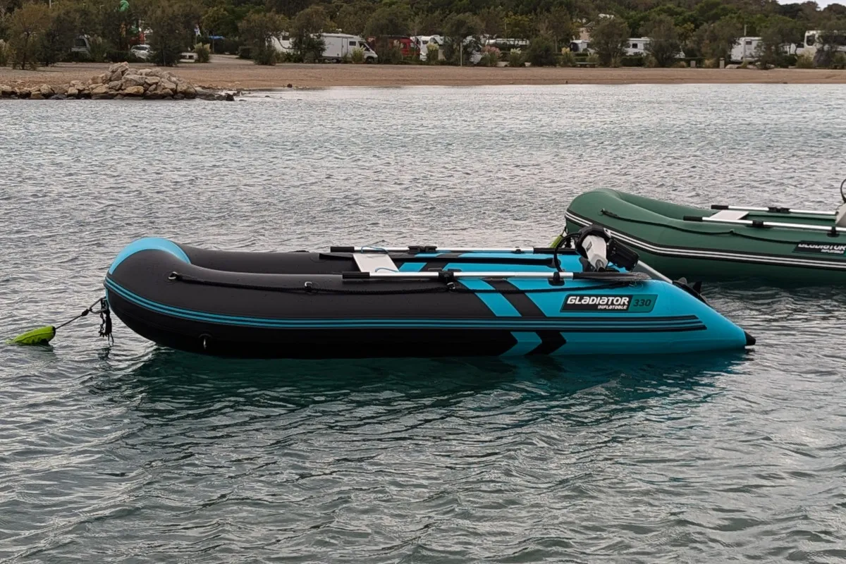 Niebiesko-czarny ponton na ryby Gladiator 330 unosi się na wodzie obok zielonej łodzi.