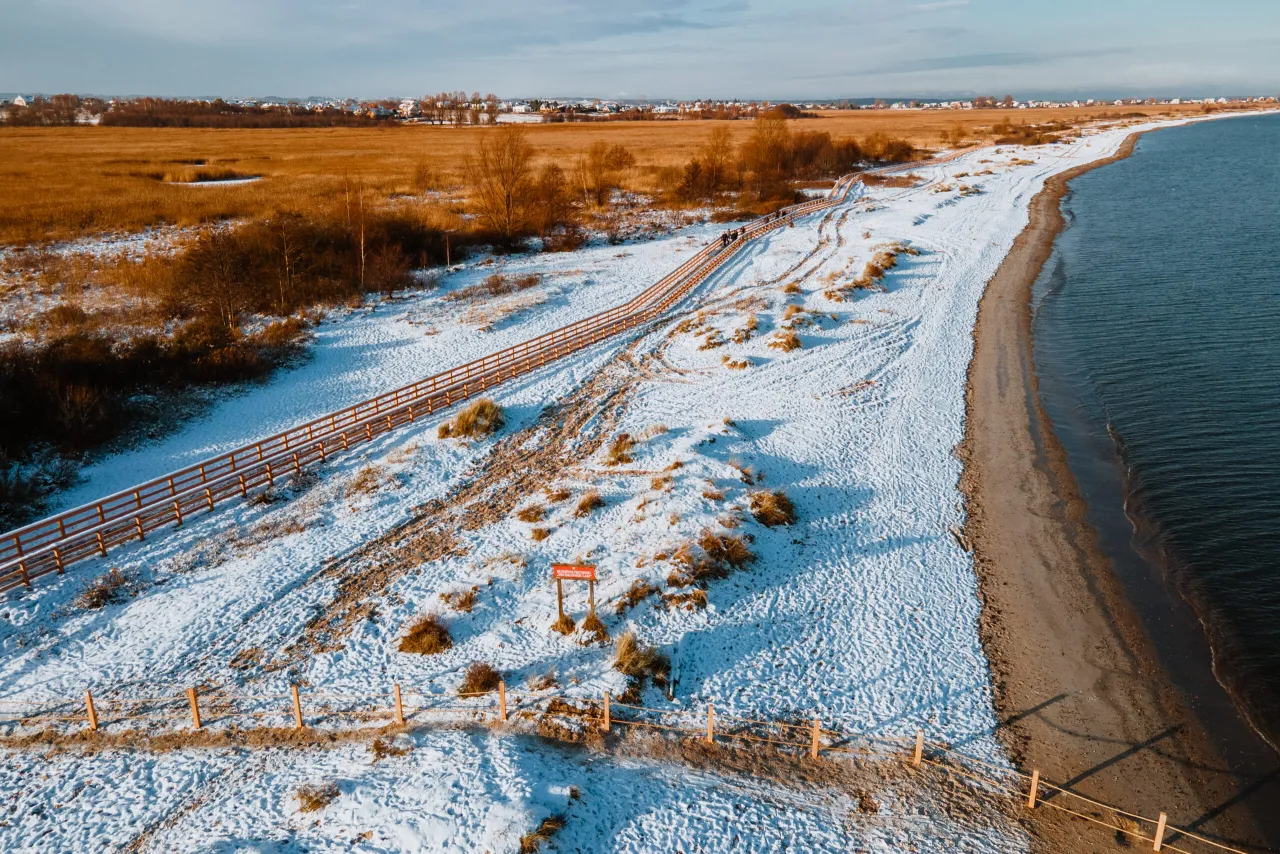 Zimowy krajobraz na rezerwacie przyrody Mechelińskie Łąki. Śnieg pokrywa plażę i wydmy, a drewniana kładka prowadzi wzdłuż wybrzeża.