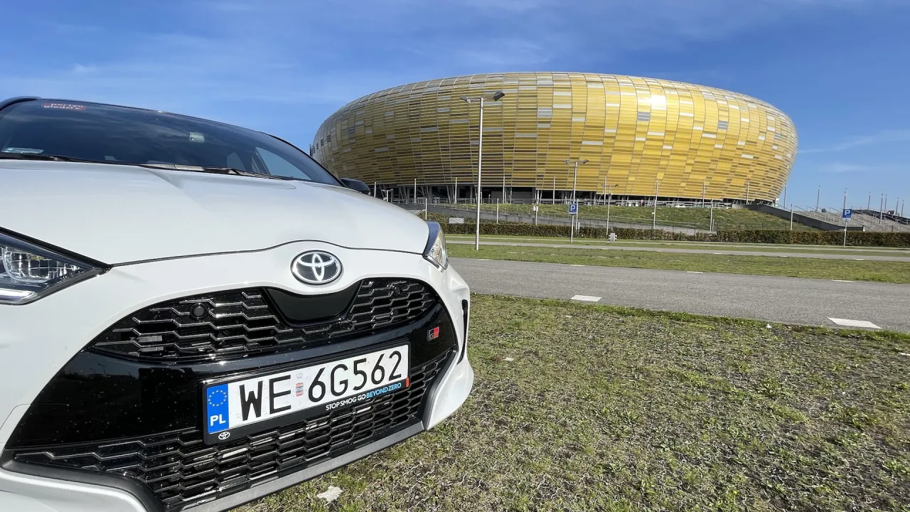 Biała Toyota Yaris Hybrid GR Sport przed żółtym stadionem. Sprawdzamy, ile pali Toyota Yaris Hybrid.