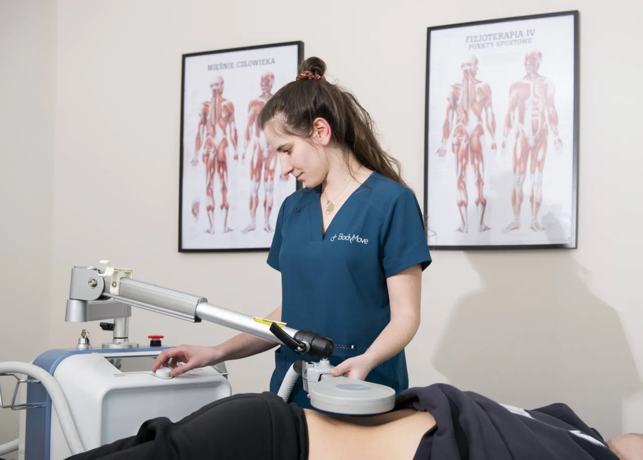 Fizjoterapeuta używa specjalistycznego sprzętu do terapii. Na ścianie wiszą anatomiczne plakaty.