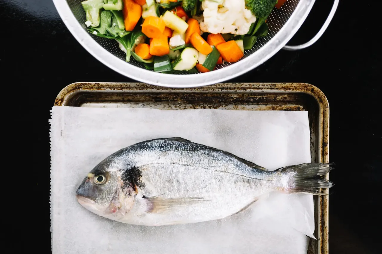Cała ryba na blasze, obok warzywa do gotowania na parze. Dowiedz się, ile gotować rybę na parze, by była idealna.