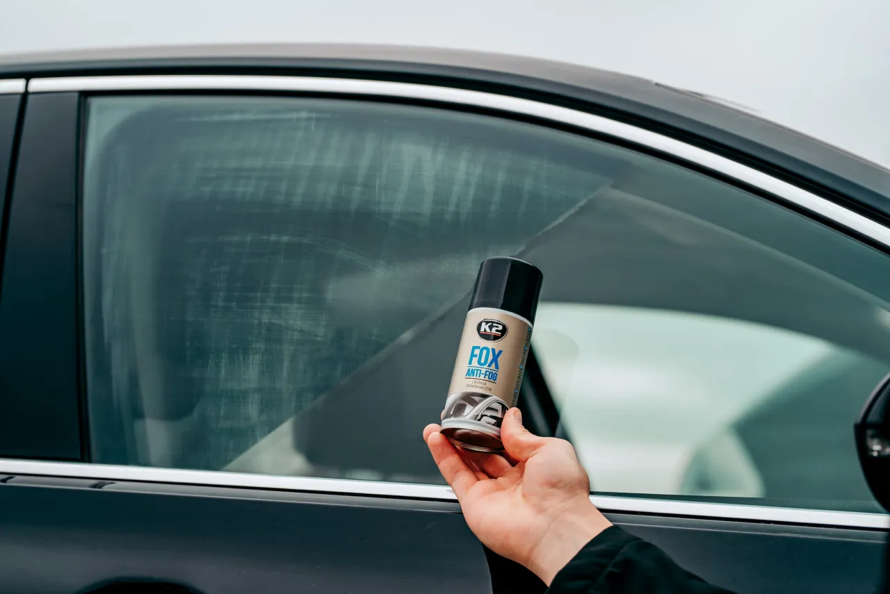 Dłoń trzyma puszkę preparatu K2 FOX ANTI-FOG obok zaparowanej szyby samochodu, by zapobiec zaparowaniu od zimnego powietrza z nawiewu.