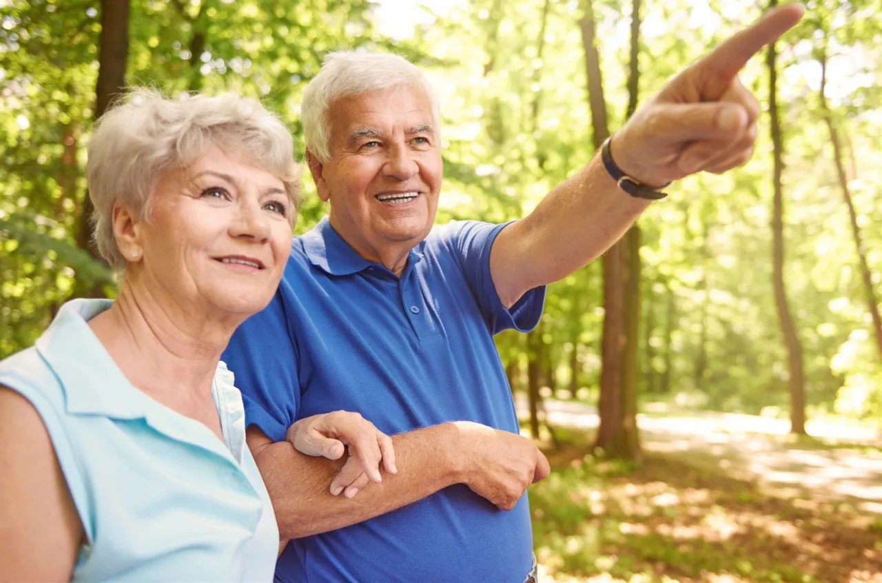 Uśmiechnięci seniorzy spacerują po lesie. Czy 50 lat to senior? On wskazuje coś palcem, ona patrzy z zaciekawieniem.