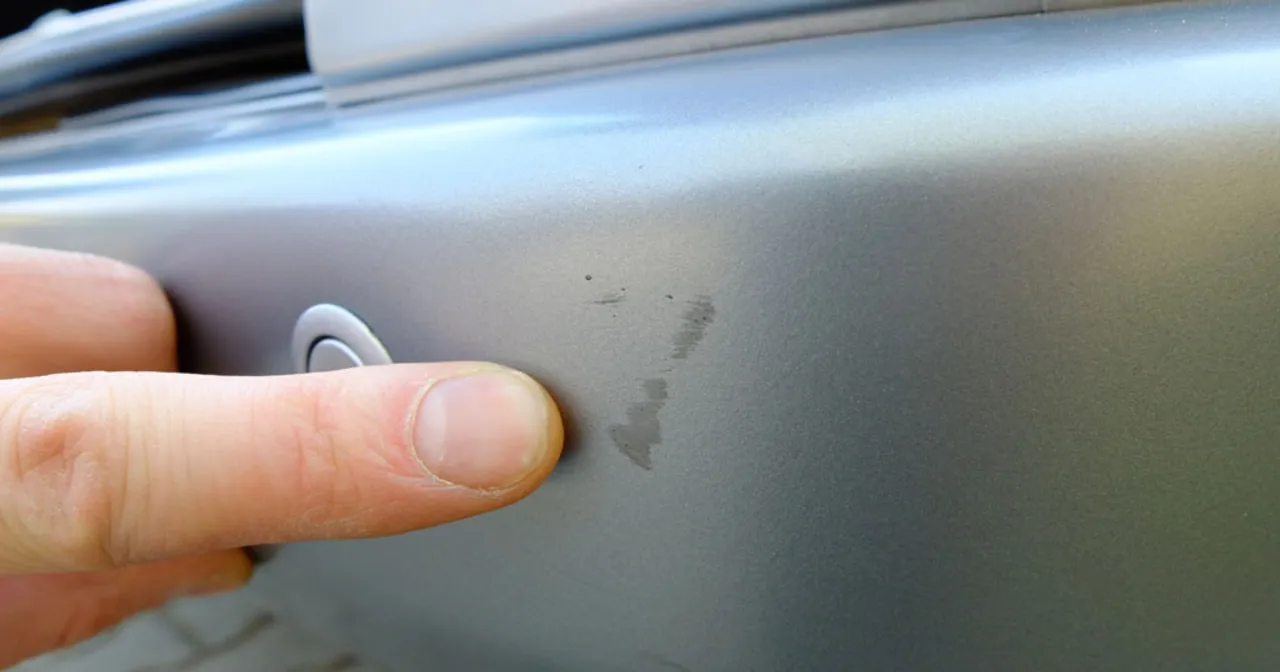 Palec wskazuje zarysowanie auta na parkingu.