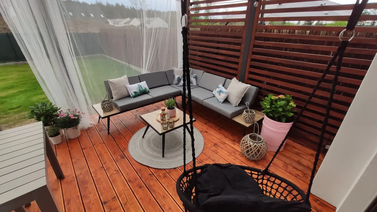 Drewniana pergola tarasowa jak zrobić? Zobacz aranżację z szarą sofą, stolikiem i huśtawką.