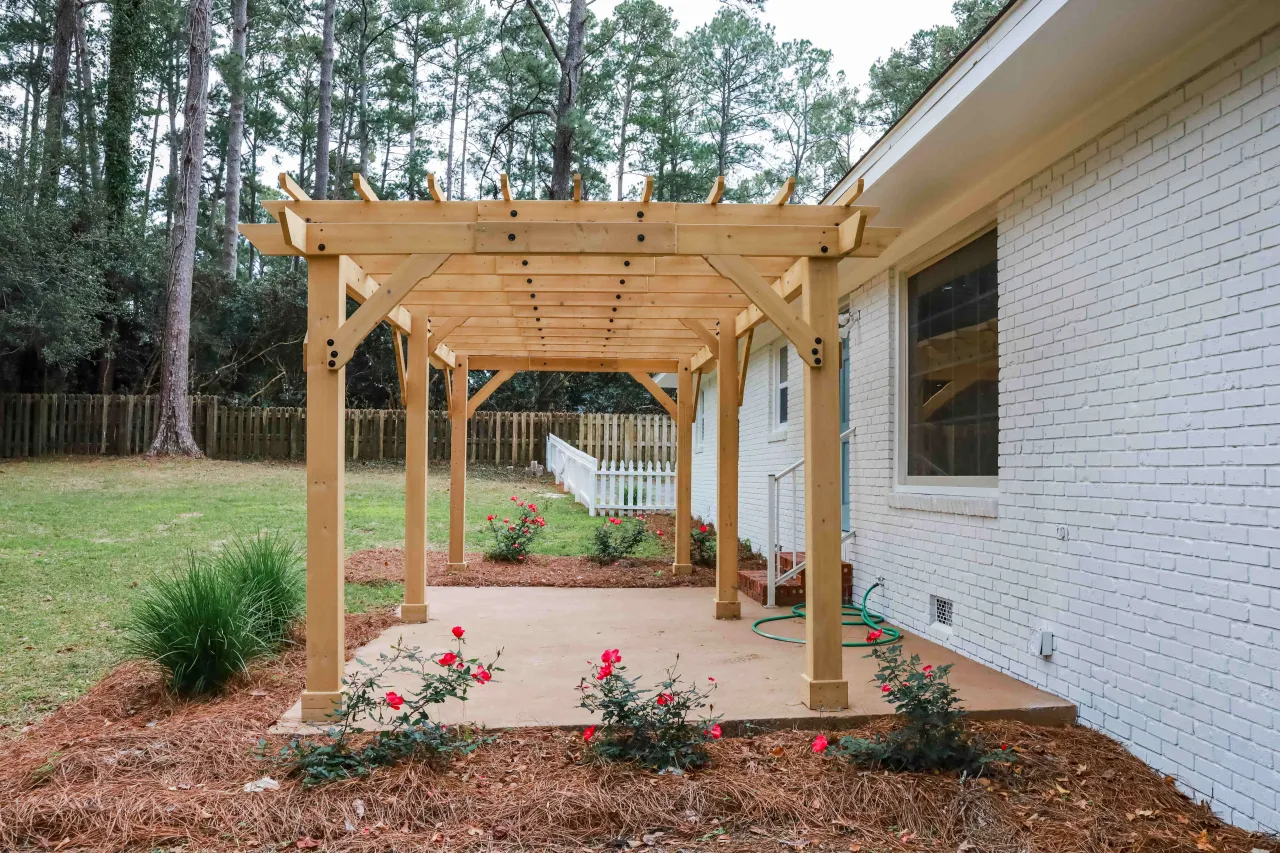 Drewniana pergola nad patio, idealna inspiracja, jak zrobić pergolę. Wokół kwitnące róże i zielony trawnik.