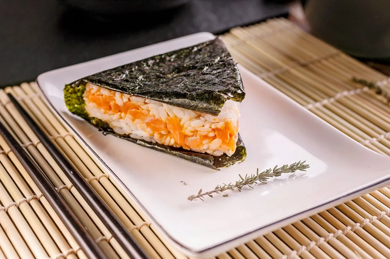 Tr&oacute;jkątne onigiri z łososiem, owinięte w nori, na białym talerzu z gałązką rozmarynu. Obok pałeczki i mata bambusowa.