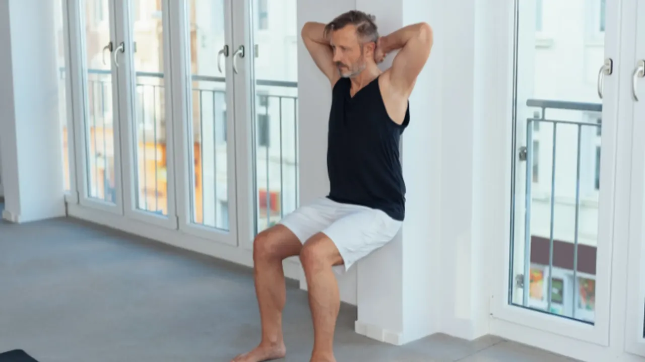 Mężczyzna wykonuje przysiad przy ścianie, zastanawiając się, czy lepszy jest trening rano czy wieczorem.