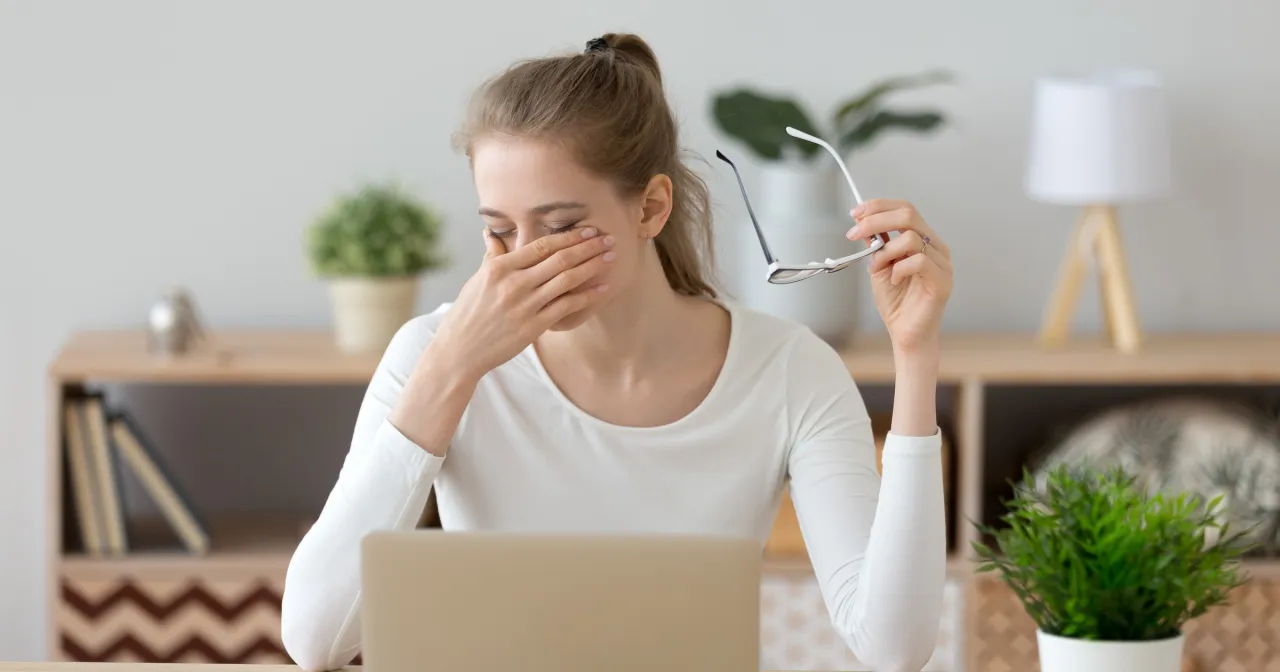 Zmęczona kobieta z okularami w dłoni pociera oczy. Brak zbieżności objawy może być spowodowany długą pracą przy komputerze.
