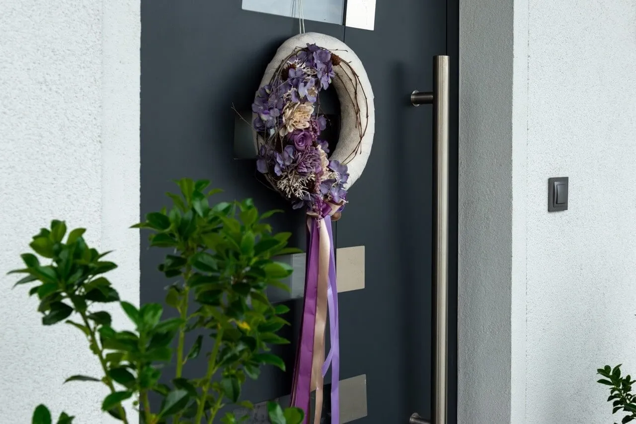 Wieniec z fioletowych kwiat&oacute;w na ciemnych drzwiach wejściowych, dodający uroku i stanowiący uzasadnienie wymiany drzwi wejściowych na bardziej estetyczne.