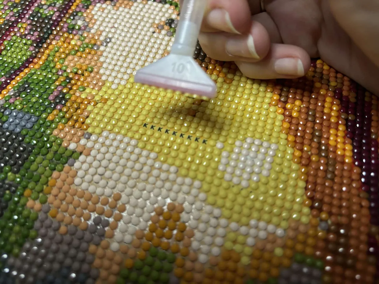 Tworzenie haftu diamentowego krok po kroku. Dłoń z aplikatorem nakłada kolorowe kryształki na pł&oacute;tno.