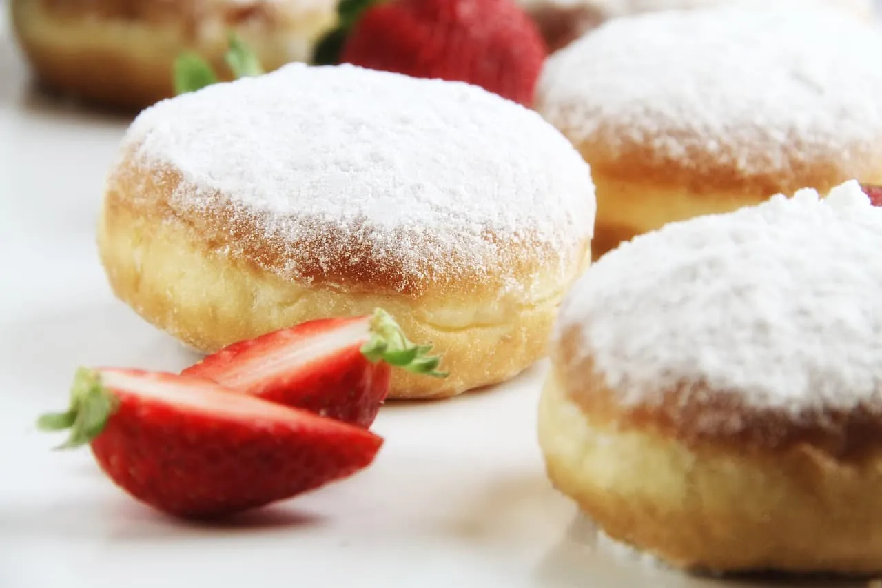 Pyszne pączki bezglutenowe posypane cukrem pudrem, obok świeże truskawki. Idealne na tłusty czwartek, oto pączki bezglutenowe przepis.