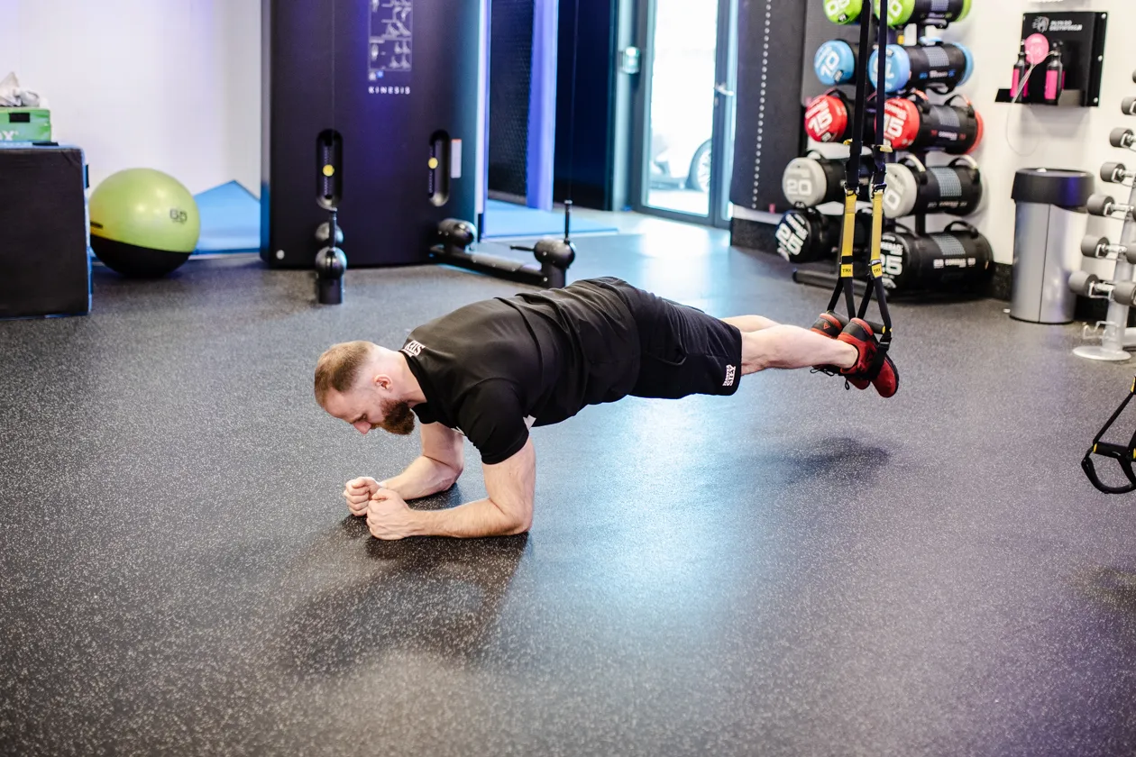 Mężczyzna wykonuje ćwiczenia na TRX, podpierając się na przedramionach i stopach zawieszonych w taśmach.
