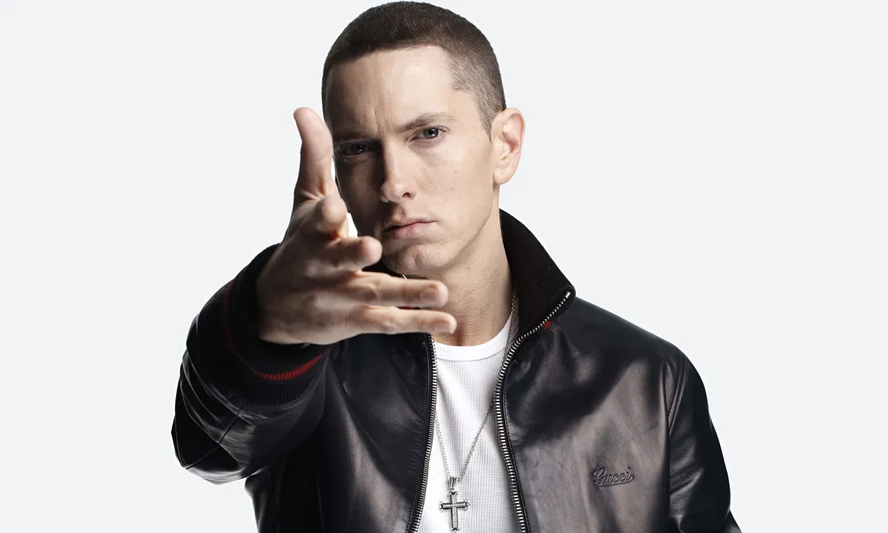 Eminem w czarnej kurtce i białej koszulce, z krzyżem na szyi, wyciąga rękę. Choć nie widać tego na zdjęciu, wiele osób zastanawia się, ile Eminem ma wzrostu.