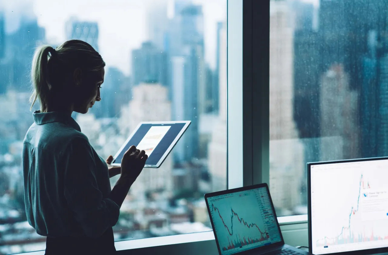 Kobieta analizuje wykresy na tablecie, podczas gdy na laptopie i monitorze widać dane z giełdy. To pokazuje, co to jest giełda &ndash; miejsce, gdzie śledzi się kursy akcji.