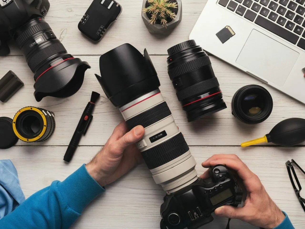 Ręce trzymają aparat z teleobiektywem, obok leżą inne obiektywy, laptop i akcesoria. Zastanawiasz się, gdzie sprzedać aparat fotograficzny?