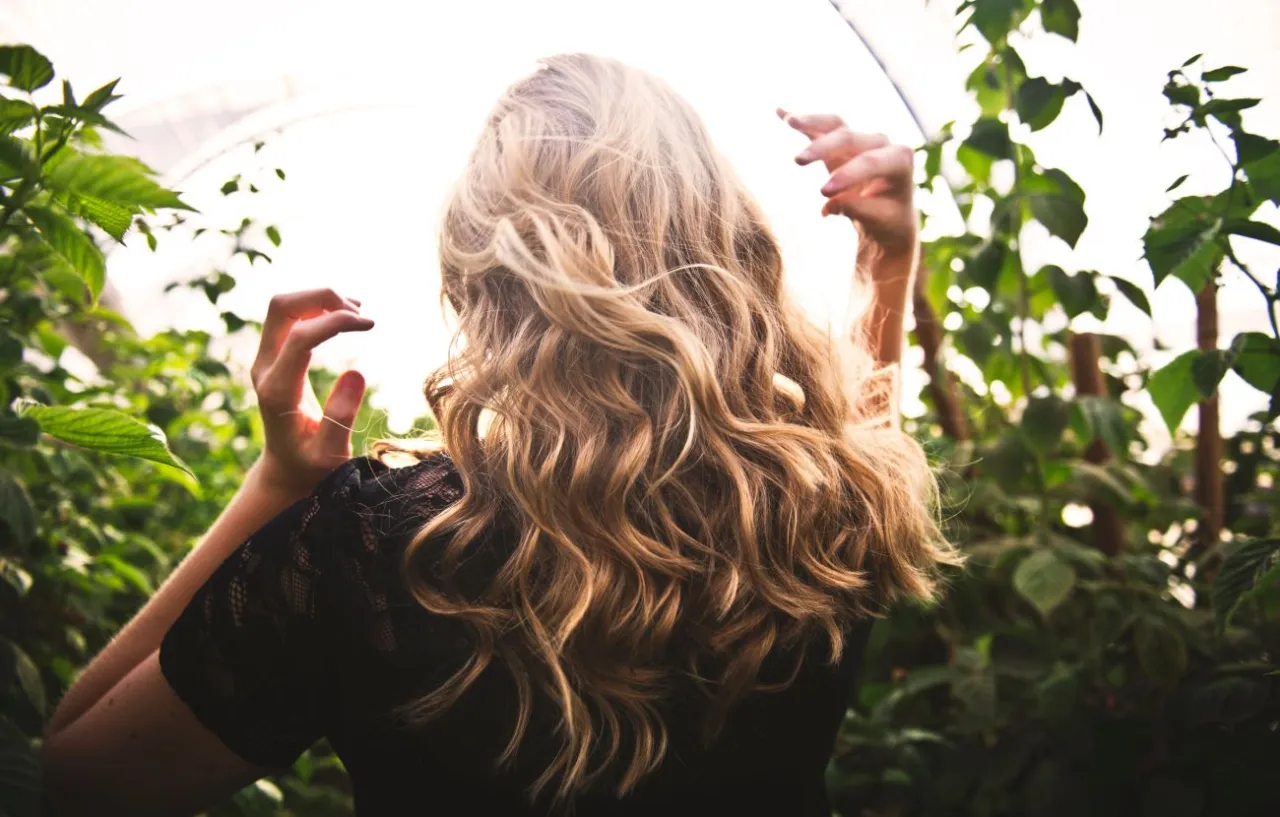 Blondynka z falowanymi włosami, jak zrobić loczki prostownicą. Słońce przebija się przez liście, tworząc magiczny efekt.