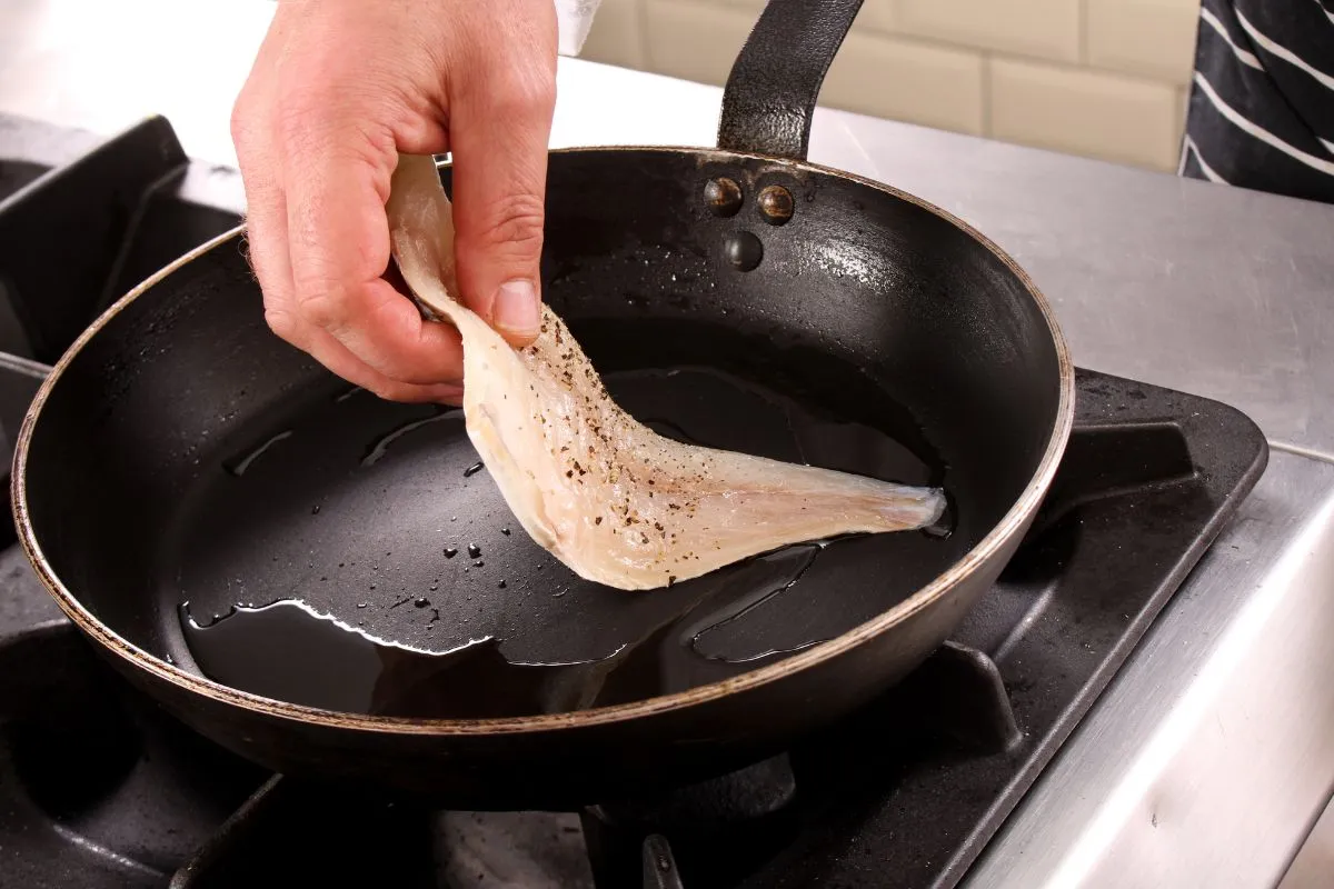 Dłoń wkłada kawałek ryby do rozgrzanej patelni z olejem. Tak zaczyna się proces, jak smażyć ryby mrożone.