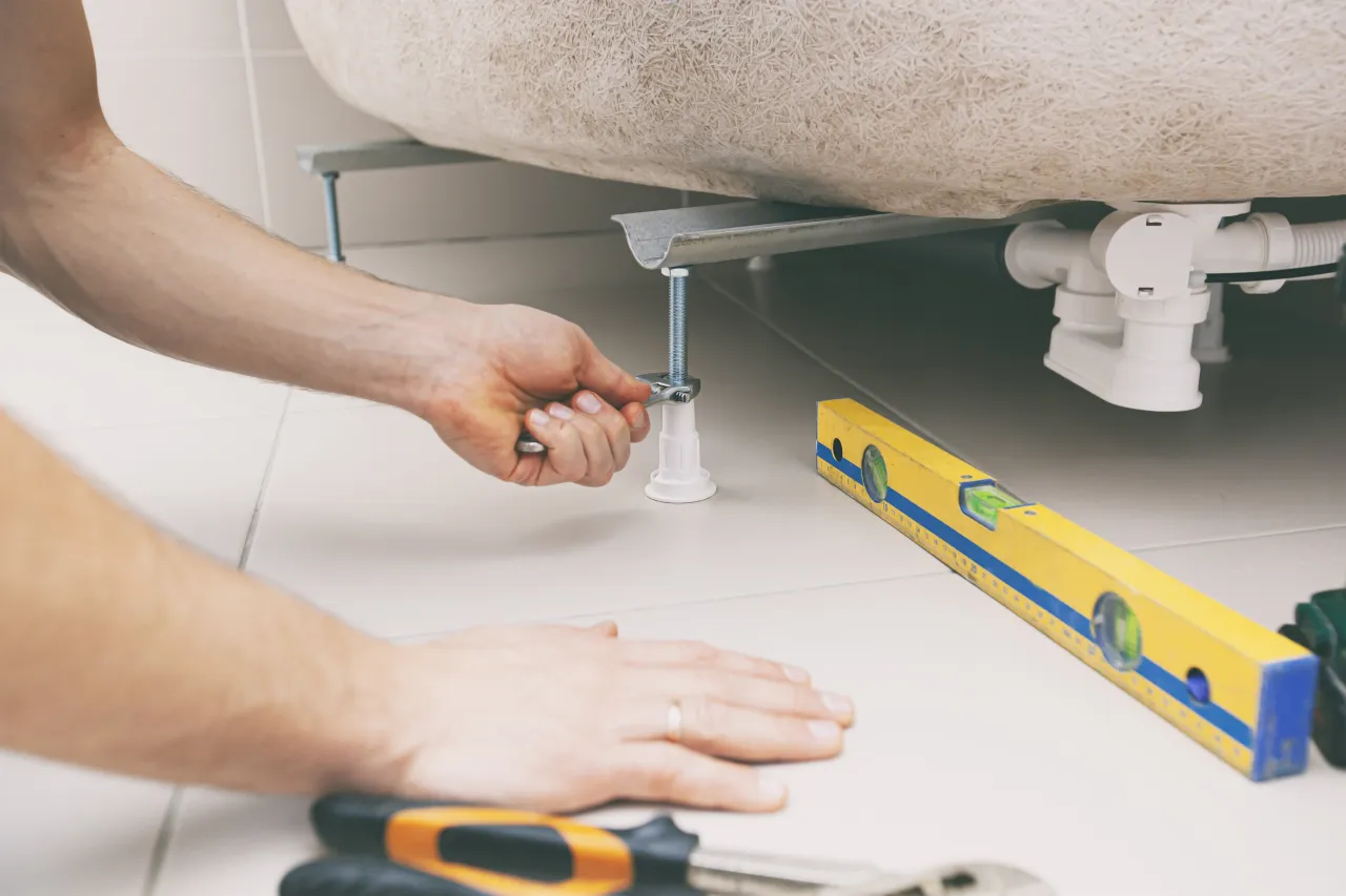 Ręka dokręca śrubę wspornika, by prawidłowo jak zamontować brodzik wysoki. Obok poziomica i narzędzia.