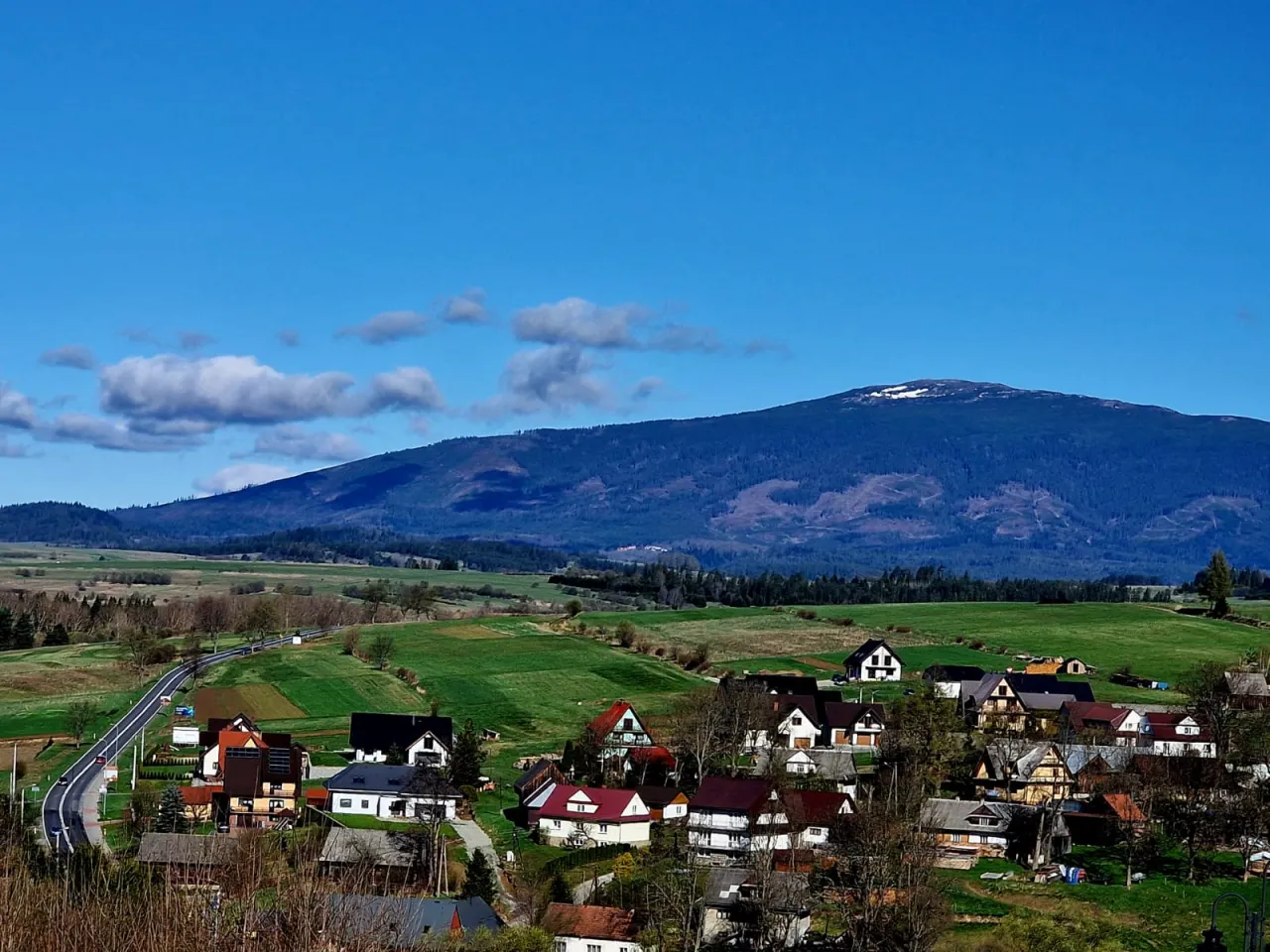 Widok na wieś u podnóża Babiej Góry. W oddali majaczy szczyt góry, a na pierwszym planie widać domy i zielone pola. Idealne miejsce na babia góra trails.