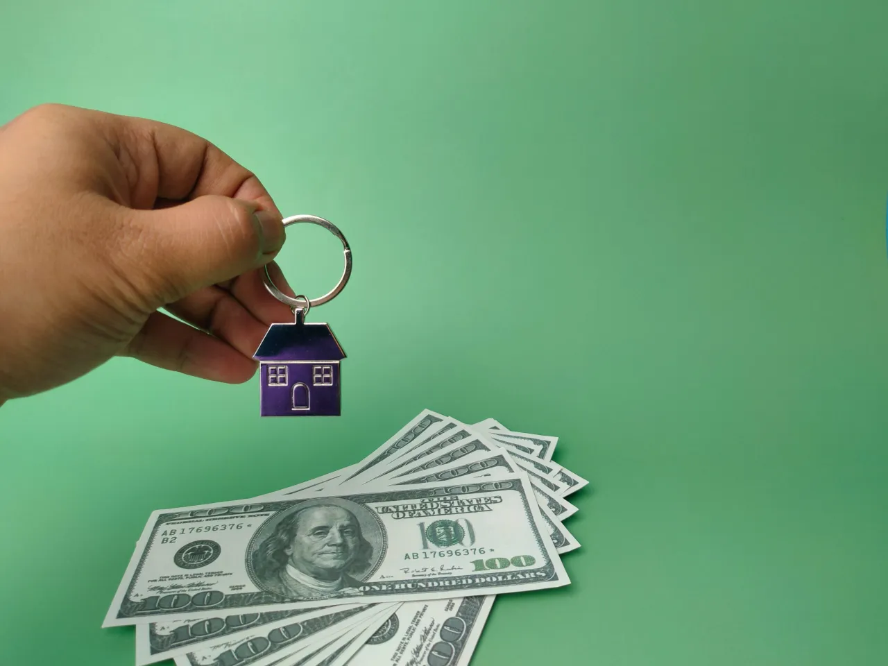 Dłoń trzyma breloczek w kształcie domu, obok leżą banknoty 100 dolarów. Zastanawiasz się, ile kosztuje wynajem mieszkania w Niemczech?